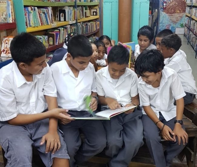 Four seated boys in school uniform gathered in foreground reading a book with several children behind them