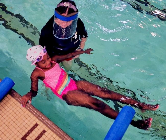 A swimming pool with a child in a pink swimsuit floating while supported by an instructor in a blue tshirt
