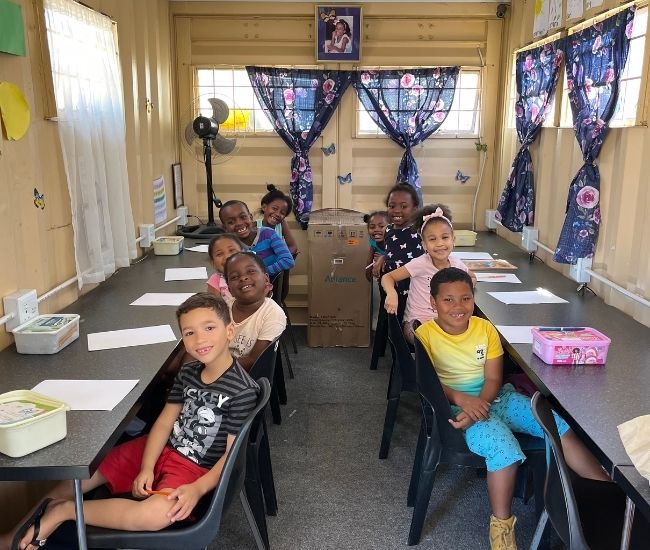 9 smiling children sitting on chairs in front of long tables on each side of a small room